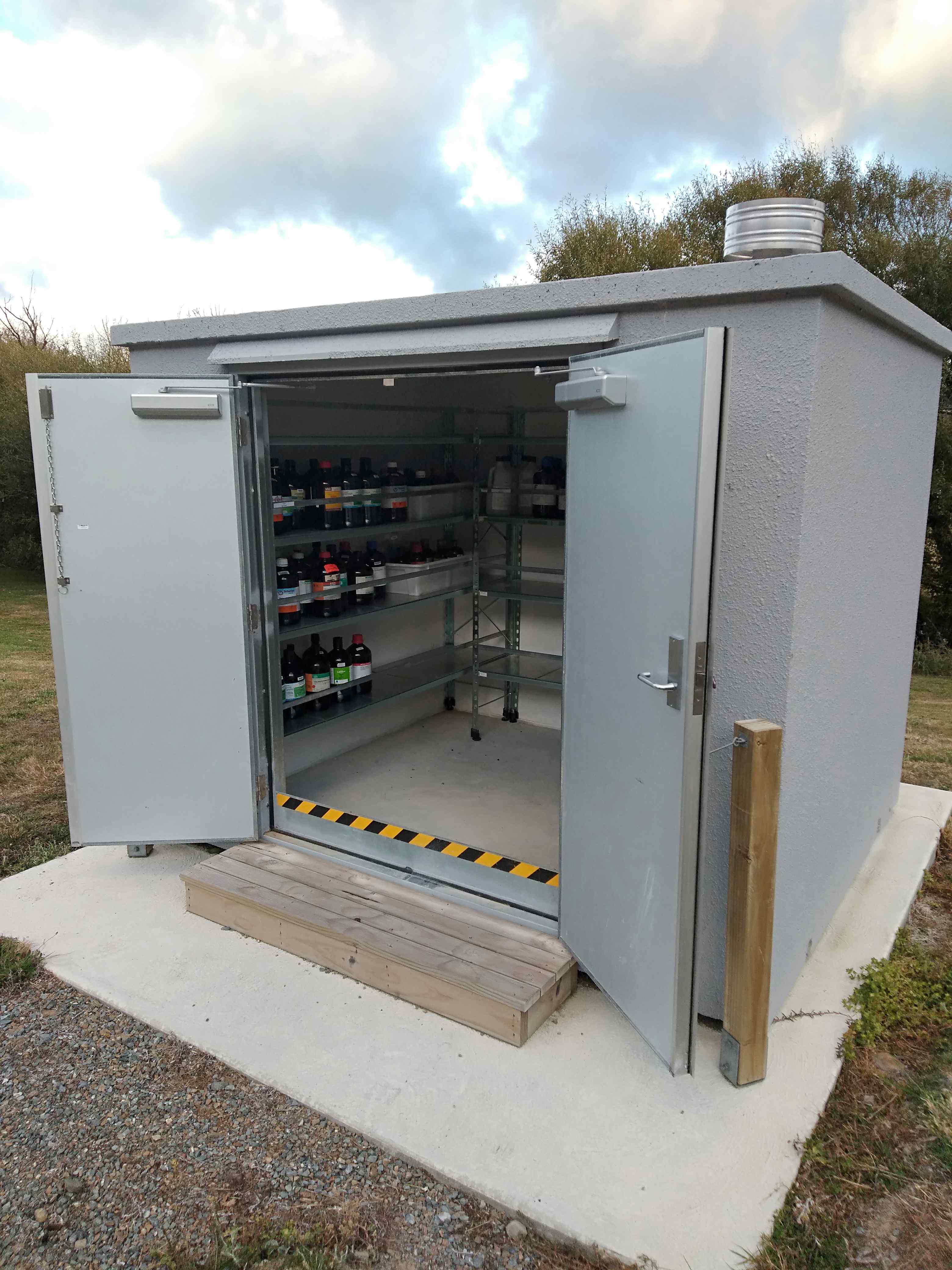 Concrete DG store with double doors open, showing shelving stocked with chemical containers inside